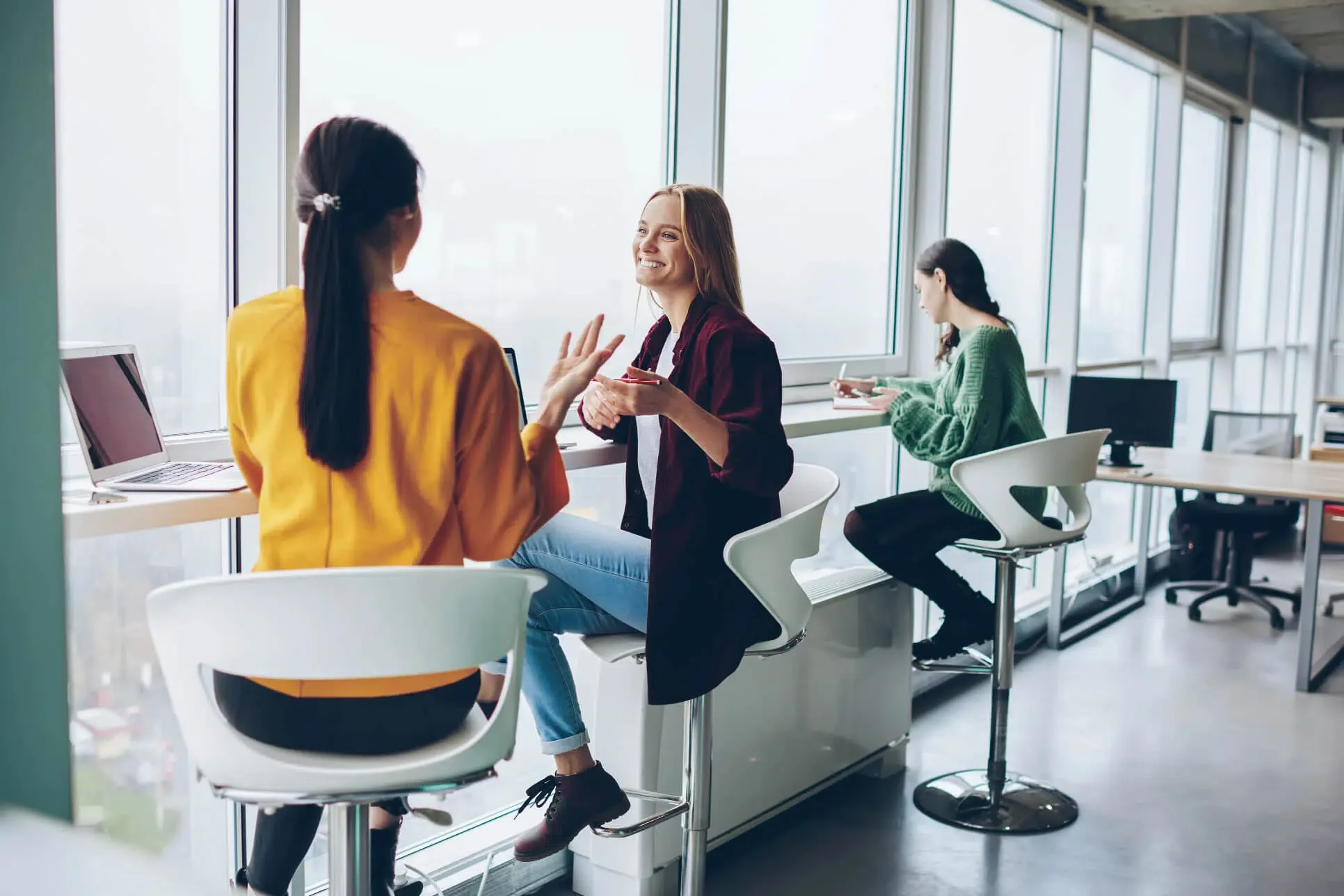 Zwei Frauen unterhalten sich lachend an einem Fenstertisch im Büro, im Hintergrund schreibt eine dritte Frau.